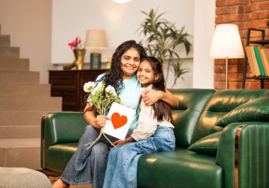 Hintli Asyalı genç anne, küçük kızına kanepede sarılıp, kalpten bir Anneler Günü 'nü kutlarken, kızının yaptığı çiçekler ve hediyelere bakarken aile bağlarını sergiliyor, sevgisini sergiliyor.