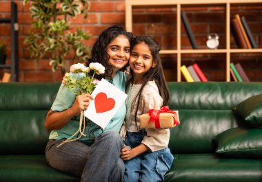 Hintli Asyalı genç anne, küçük kızına kanepede sarılıp, kalpten bir Anneler Günü 'nü kutlarken, kızının yaptığı çiçekler ve hediyelere bakarken aile bağlarını sergiliyor, sevgisini sergiliyor.