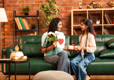 Hintli Asyalı genç anne, küçük kızına kanepede sarılıp, kalpten bir Anneler Günü 'nü kutlarken, kızının yaptığı çiçekler ve hediyelere bakarken aile bağlarını sergiliyor, sevgisini sergiliyor.