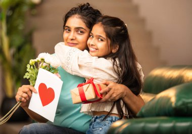 Hintli Asyalı genç anne, küçük kızına kanepede sarılıp, kalpten bir Anneler Günü 'nü kutlarken, kızının yaptığı çiçekler ve hediyelere bakarken aile bağlarını sergiliyor, sevgisini sergiliyor.
