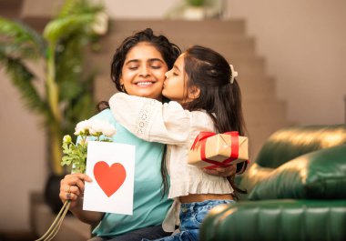 Hintli Asyalı genç anne, küçük kızına kanepede sarılıp, kalpten bir Anneler Günü 'nü kutlarken, kızının yaptığı çiçekler ve hediyelere bakarken aile bağlarını sergiliyor, sevgisini sergiliyor.