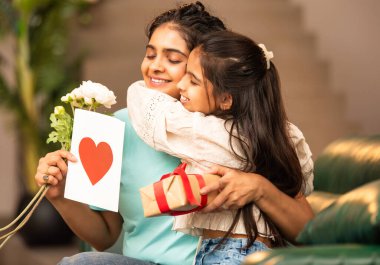 Hintli Asyalı genç anne, küçük kızına kanepede sarılıp, kalpten bir Anneler Günü 'nü kutlarken, kızının yaptığı çiçekler ve hediyelere bakarken aile bağlarını sergiliyor, sevgisini sergiliyor.