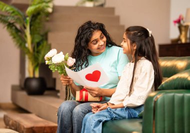 Hintli Asyalı genç anne, küçük kızına kanepede sarılıp, kalpten bir Anneler Günü 'nü kutlarken, kızının yaptığı çiçekler ve hediyelere bakarken aile bağlarını sergiliyor, sevgisini sergiliyor.