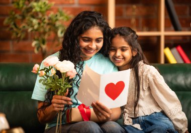 Hintli Asyalı genç anne, küçük kızına kanepede sarılıp, kalpten bir Anneler Günü 'nü kutlarken, kızının yaptığı çiçekler ve hediyelere bakarken aile bağlarını sergiliyor, sevgisini sergiliyor.