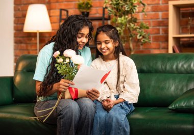 Hintli Asyalı genç anne, küçük kızına kanepede sarılıp, kalpten bir Anneler Günü 'nü kutlarken, kızının yaptığı çiçekler ve hediyelere bakarken aile bağlarını sergiliyor, sevgisini sergiliyor.