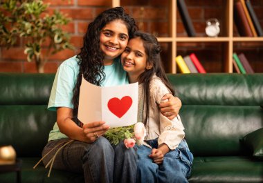 Hintli Asyalı genç anne, küçük kızına kanepede sarılıp, kalpten bir Anneler Günü 'nü kutlarken, kızının yaptığı çiçekler ve hediyelere bakarken aile bağlarını sergiliyor, sevgisini sergiliyor.