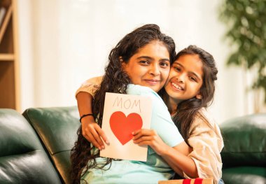 Hintli Asyalı genç anne, küçük kızına kanepede sarılıp, kalpten bir Anneler Günü 'nü kutlarken, kızının yaptığı çiçekler ve hediyelere bakarken aile bağlarını sergiliyor, sevgisini sergiliyor.