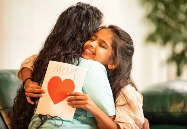 Hintli Asyalı genç anne, küçük kızına kanepede sarılıp, kalpten bir Anneler Günü 'nü kutlarken, kızının yaptığı çiçekler ve hediyelere bakarken aile bağlarını sergiliyor, sevgisini sergiliyor.