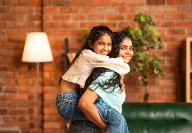 Hindistanlı şirin küçük kız, modern bir oturma odasında annesinin sırtında neşe içinde gülüyor ve eğleniyor. Sevgi dolu mutlu bir aile anı yaratıyor.