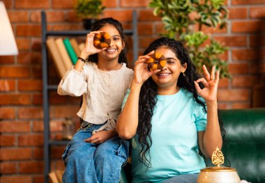 Hintli Asyalı genç bir anne ve şirin küçük bir kız kanepede oturuyor modern bir evde, neşeyle yuvarlak bir çörek tutuyor, delikten bakıyor, gülümsüyor ve iyi bir el hareketi yapıyor.