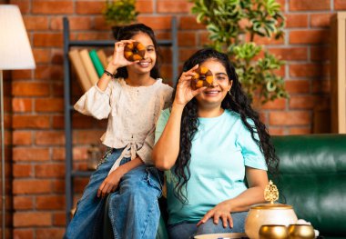 Hintli Asyalı genç bir anne ve şirin küçük bir kız kanepede oturuyor modern bir evde, neşeyle yuvarlak bir çörek tutuyor, delikten bakıyor, gülümsüyor ve iyi bir el hareketi yapıyor.