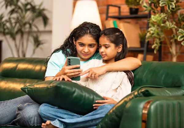 Hintli Asyalı genç anne kız evdeki rahat modern koltukta oturuyor, akıllı telefonu online alışveriş için kullanıyor, interneti tarıyor, sosyal medyayı karıştırıyor, video görüşmeleri yapıyor, dijital yaşam tarzının tadını çıkarıyor.