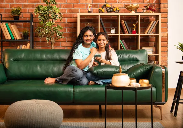 Hintli Asyalı genç anne kız evdeki rahat modern koltukta oturuyor, akıllı telefonu online alışveriş için kullanıyor, interneti tarıyor, sosyal medyayı karıştırıyor, video görüşmeleri yapıyor, dijital yaşam tarzının tadını çıkarıyor.