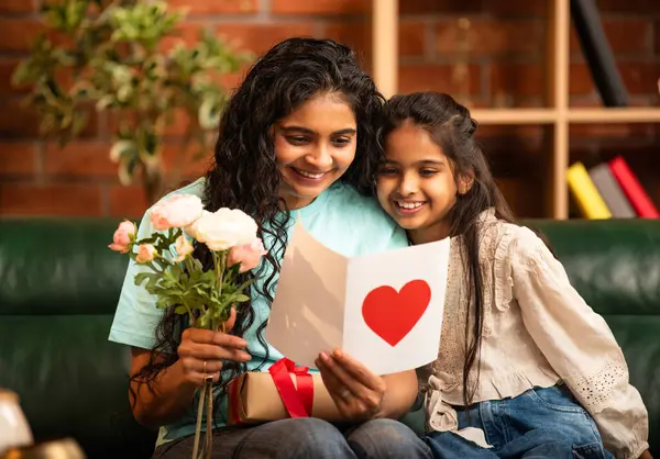 Hintli Asyalı genç anne, küçük kızına kanepede sarılıp, kalpten bir Anneler Günü 'nü kutlarken, kızının yaptığı çiçekler ve hediyelere bakarken aile bağlarını sergiliyor, sevgisini sergiliyor.