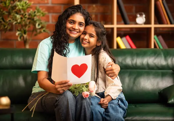Hintli Asyalı genç anne, küçük kızına kanepede sarılıp, kalpten bir Anneler Günü 'nü kutlarken, kızının yaptığı çiçekler ve hediyelere bakarken aile bağlarını sergiliyor, sevgisini sergiliyor.