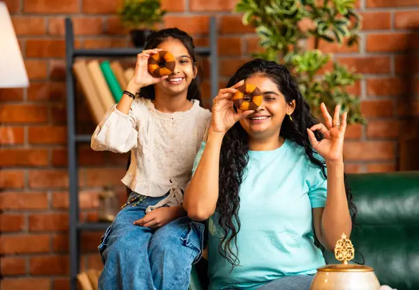 Hintli Asyalı genç bir anne ve şirin küçük bir kız kanepede oturuyor modern bir evde, neşeyle yuvarlak bir çörek tutuyor, delikten bakıyor, gülümsüyor ve iyi bir el hareketi yapıyor.