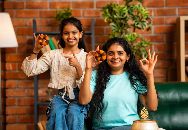 Hintli Asyalı genç bir anne ve şirin küçük bir kız kanepede oturuyor modern bir evde, neşeyle yuvarlak bir çörek tutuyor, delikten bakıyor, gülümsüyor ve iyi bir el hareketi yapıyor.