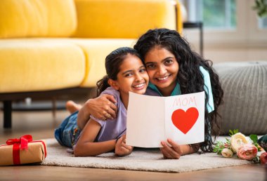 Hintli Asyalı genç anne ve küçük kızı modern şık bir evde, halının üzerinde uzanmış neşeli bir Anneler Günü kartı okuyorlar. Aile bağlarını paylaşıyorlar.