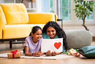 Hintli Asyalı genç anne ve küçük kızı modern şık bir evde, halının üzerinde uzanmış neşeli bir Anneler Günü kartı okuyorlar. Aile bağlarını paylaşıyorlar.