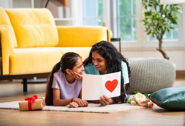 Hintli Asyalı genç anne ve küçük kızı modern şık bir evde, halının üzerinde uzanmış neşeli bir Anneler Günü kartı okuyorlar. Aile bağlarını paylaşıyorlar.