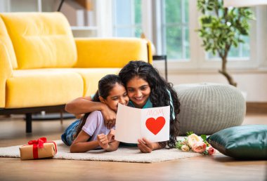 Hintli Asyalı genç anne ve küçük kızı modern şık bir evde, halının üzerinde uzanmış neşeli bir Anneler Günü kartı okuyorlar. Aile bağlarını paylaşıyorlar.