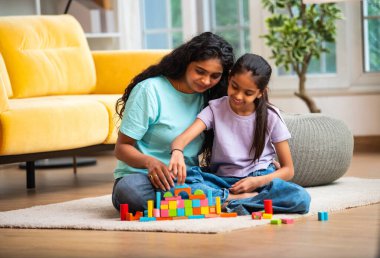 Hint Asyalı genç anne ve küçük kızı renkli ahşap oyuncaklar kullanarak ev inşa ederken modern müsrif bir evin halılı zemininde birlikte oturuyorlar ve kapalı alanda kaliteli zaman geçiriyorlar.