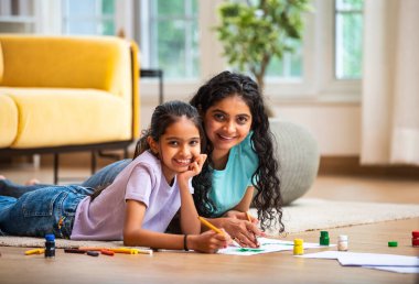 Hintli Asyalı genç anne ve küçük kızı modern bir evin zemininde uzanmış, kağıt üzerinde fırça ve su renklerini kullanarak sanat, çizim ve resim faaliyetleriyle mutlu mesut ilgileniyorlar.