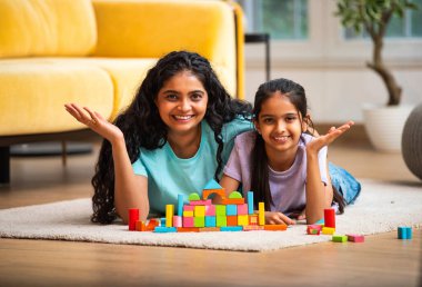 Hint Asyalı genç anne ve küçük kızı renkli ahşap oyuncaklar kullanarak ev inşa ederken modern müsrif bir evin halılı zemininde birlikte oturuyorlar ve kapalı alanda kaliteli zaman geçiriyorlar.