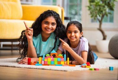 Hint Asyalı genç anne ve küçük kızı renkli ahşap oyuncaklar kullanarak ev inşa ederken modern müsrif bir evin halılı zemininde birlikte oturuyorlar ve kapalı alanda kaliteli zaman geçiriyorlar.