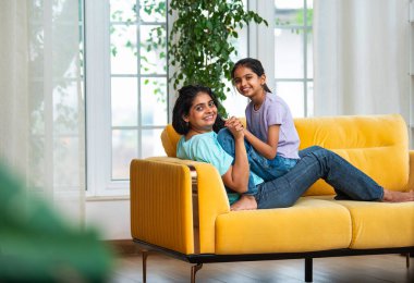 Hintli Asyalı genç anne küçük kızıyla modern oturma odasındaki kanepede neşeyle tahterevalli gibi oynuyor, eğlenceli bağlılık anları, mutluluk, kahkaha ve kapalı alanda kaliteli zaman geçirmenin tadını çıkarıyor.