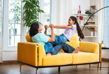 Hintli Asyalı genç anne küçük kızıyla modern oturma odasındaki kanepede neşeyle tahterevalli gibi oynuyor, eğlenceli bağlılık anları, mutluluk, kahkaha ve kapalı alanda kaliteli zaman geçirmenin tadını çıkarıyor.