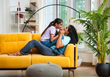 Hintli Asyalı genç anne küçük kızıyla modern oturma odasındaki kanepede neşeyle tahterevalli gibi oynuyor, eğlenceli bağlılık anları, mutluluk, kahkaha ve kapalı alanda kaliteli zaman geçirmenin tadını çıkarıyor.