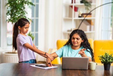 Hintli Asyalı genç anne, modern evdeki dizüstü bilgisayarında çalışırken, sevimli küçük kızı dikkat çekmek için elini çekerek, iş-yaşam dengesi mücadelesi sergiliyor.