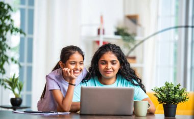 Hintli Asyalı genç anne bilgisayarının başında uzaktan bilgisayarla çalışırken tapılası küçük kızı ona arkadan sarılıyor ve çalışmalarını modern, havadar, şık bir evde izliyor.