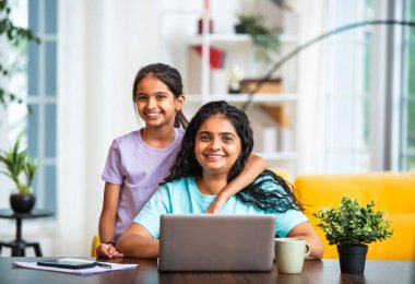 Hintli Asyalı genç anne bilgisayarının başında uzaktan bilgisayarla çalışırken tapılası küçük kızı ona arkadan sarılıyor ve çalışmalarını modern, havadar, şık bir evde izliyor.