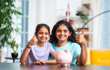 Hintli Asyalı genç bir kadın ve şirin küçük bir kız birlikte para biriktiriyorlar, modern evdeki masa başındaki pembe domuzcuk bankasına para koyuyorlar, erken tasarruf alışkanlıklarını, disiplini, finansal okur yazarlığı öğretiyorlar.