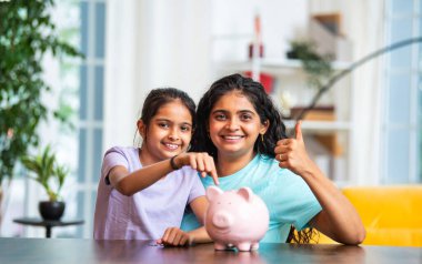 Hintli Asyalı genç bir kadın ve şirin küçük bir kız birlikte para biriktiriyorlar, modern evdeki masa başındaki pembe domuzcuk bankasına para koyuyorlar, erken tasarruf alışkanlıklarını, disiplini, finansal okur yazarlığı öğretiyorlar.