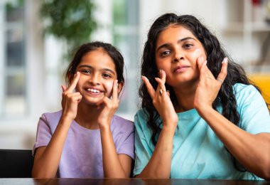 Hintli Asyalı genç bir kadın ve sevimli küçük bir kız mutlu ve şakacı bir şekilde yanaklarına krem sürerken evde birlikte aynaya bakıp eğleniyorlar.