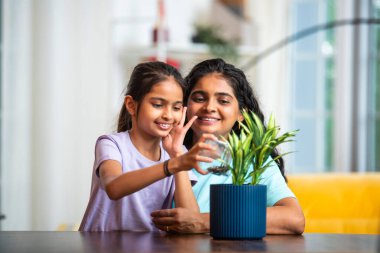 Hintli Asyalı genç bir kadın ve şirin küçük kızı yeşil saksı bitkisine bozuk para koyarken modern bir evde masa başında, finansal büyümeyi ve erken tasarruf alışkanlıklarını simgeliyor.