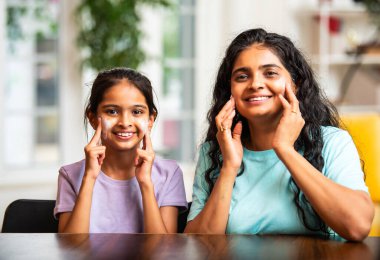 Hintli Asyalı genç bir kadın ve sevimli küçük bir kız mutlu ve şakacı bir şekilde yanaklarına krem sürerken evde birlikte aynaya bakıp eğleniyorlar.