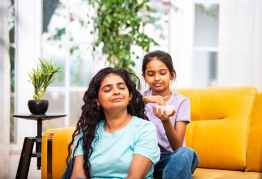 Hindistanlı şirin küçük kız, genç annesine yumuşak bir kafa masajı yapıyor, evdeki havadar, modern bir oturma odasında rahat bir kanepede birlikte otururken.