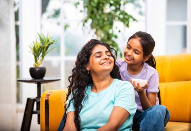 Hindistanlı şirin küçük kız, genç annesine yumuşak bir kafa masajı yapıyor, evdeki havadar, modern bir oturma odasında rahat bir kanepede birlikte otururken.