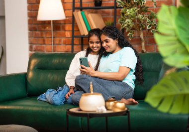 Hintli Asyalı genç anne kız evdeki rahat modern koltukta oturuyor, akıllı telefonu online alışveriş için kullanıyor, interneti tarıyor, sosyal medyayı karıştırıyor, video görüşmeleri yapıyor, dijital yaşam tarzının tadını çıkarıyor.