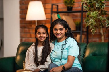 Hintli Asyalı genç anne ve tatlı küçük kızının portresi sıcak bir şekilde gülümsüyor ve doğrudan kameraya bakıyor rahat bir kanepede otururken şık, müsrif modern ev içinde.