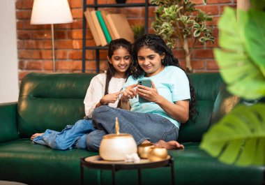 Hintli Asyalı genç anne kız evdeki rahat modern koltukta oturuyor, akıllı telefonu online alışveriş için kullanıyor, interneti tarıyor, sosyal medyayı karıştırıyor, video görüşmeleri yapıyor, dijital yaşam tarzının tadını çıkarıyor.