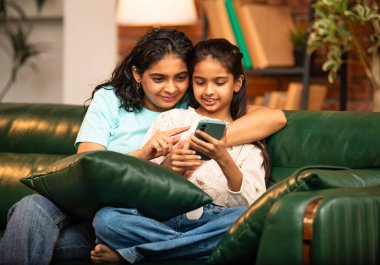 Hintli Asyalı genç anne kız evdeki rahat modern koltukta oturuyor, akıllı telefonu online alışveriş için kullanıyor, interneti tarıyor, sosyal medyayı karıştırıyor, video görüşmeleri yapıyor, dijital yaşam tarzının tadını çıkarıyor.