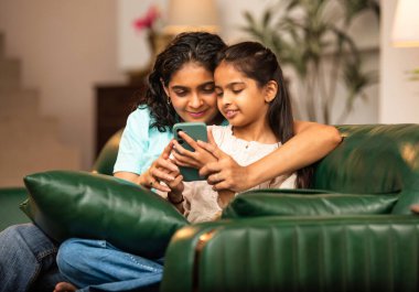 Hintli Asyalı genç anne kız evdeki rahat modern koltukta oturuyor, akıllı telefonu online alışveriş için kullanıyor, interneti tarıyor, sosyal medyayı karıştırıyor, video görüşmeleri yapıyor, dijital yaşam tarzının tadını çıkarıyor.