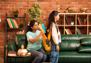 Hintli bir anne, kızının okula gitmesine yardım etmek için kitap paketliyor, okul çantasına beslenme çantası koyuyor, ona sevgiyle sarılıyor ve sıcak bir ev ortamında şefkatle onu uğurluyor.