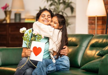 Hintli Asyalı genç anne, küçük kızına kanepede sarılıp, kalpten bir Anneler Günü 'nü kutlarken, kızının yaptığı çiçekler ve hediyelere bakarken aile bağlarını sergiliyor, sevgisini sergiliyor.