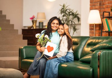 Hintli Asyalı genç anne, küçük kızına kanepede sarılıp, kalpten bir Anneler Günü 'nü kutlarken, kızının yaptığı çiçekler ve hediyelere bakarken aile bağlarını sergiliyor, sevgisini sergiliyor.